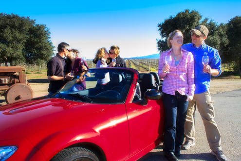 3 couples kiss around a red convertible sports car in front of a vineyard.