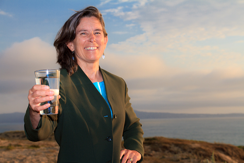 MCWD Director jan Shriner poses with a glass of water with sand dunes, Monterey Bay and Monterey Peninsula in the background.