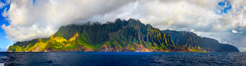 The Kauai island as seen from a boat on the ocean with bright-colored foliage and cloud-shrouded mountains.