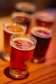 6 beer tasting glasses stagger out of focus across a wooden bar with colorful lights.