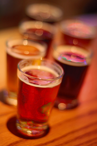 Beer tasting glasses stagger out of focus across a wooden bar with dreamy colored bar lights reflecting off the surface of the beers. 