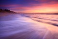 Colorfully lit surf rushes up the beach at twilight with a colorful sky over Monterey and silhouetted sand dunes.