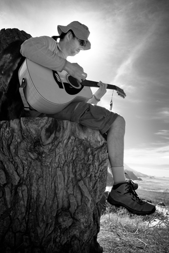 A guitar player sits on a wooden throne with feet dangling over the edge of a cliff where the Santa Lucia Mountains and Pacific Ocean collide. 