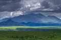 Shaded green tundra valley leading into a sunlight patch with dark blue mountains and dramatic clouds in the background.