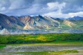 Streams from glacial melt fill a valley leading into tundra and different colored mountains with dramatic clouds.
