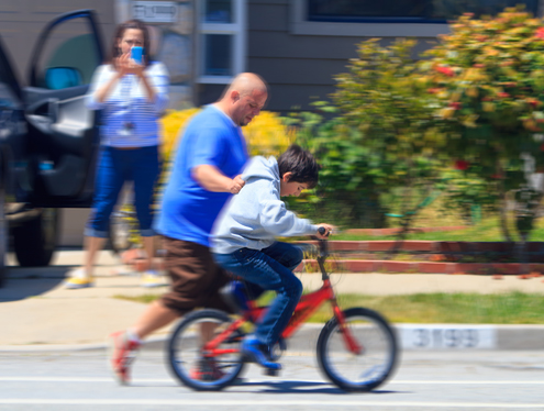 My neighbor teaching his son to ride a bike while his wife captures the moment on with her phone camera. The quality of a community might be measured by how inviting its streets are to learning to ride a bike. 