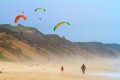 Parasailors hover above sand dunes and a beach with 2 people walking on the beach.