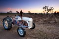 An old tractor in front of tilled fields, oak tree, and sunset.