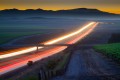 Long exposure of car lights trace the path of a highway through a vineyard landscape at dusk.