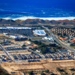 Aerial photo of half-completed development project including a theater, big box shopping center, commercial offices, medical facilities, single and multi family housing, hotel, and restaurants with the Fort Ord Dunes State Park and Monterey Bay in the background.