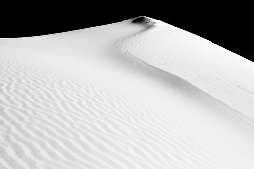 The deep blue sky goes to black with a deep red filter over the gently curving crest of a pure white sand dune. Part of a black and white portfolio of landscape and abstract nature photographs depicting Science Fiction-like imagery from distant galaxies to Earth’s prehistoric natural history. 