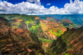Red canyons walls dressed with bright green foliage with a long white waterfall and river, blue sky, and fluffy clouds.