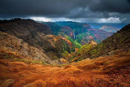 Waimea Canyon is called the Grand Canyon of the Pacific. It is a breathtaking experience standing on the edge and looking down into a kaleidoscope of colors: deep red volcanic rock and dirt, plush green forest, and long white waterfalls under a dark gray sky ready to dump and afternoon rain. 