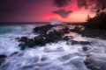 The tide draining over lava rocks towards another incoming wave crashing with a red sunset sky.