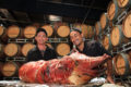 2 Chefs display a roast pig with a wall of wine barrels in the background.