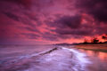 Purple, magenta, red and orange sunset reflected in the pacific ocean with high tide pouring over rocks.
