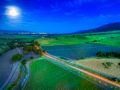 Aerial drone photo of vineyards and fields along River Road winding south through the Salinas Valley by the light of a full moon with the city lights of Gonzales and Soledad in the distance.