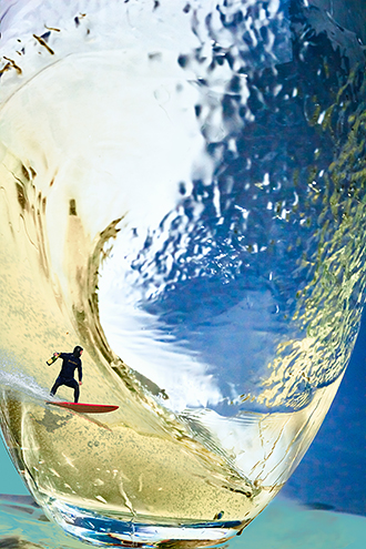 Photo illustration of a surfer from Marina State Beach with a bottle of bubbly in his hand and the cork flying off rides a wave of sparkling wine as it swooshes around in a glass. 