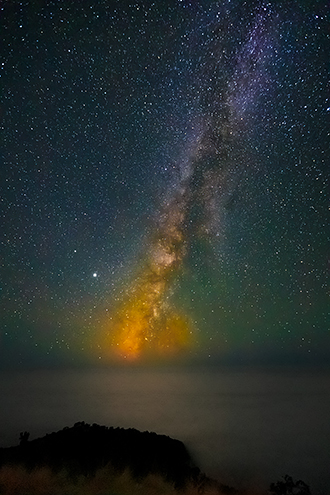 The the brightest area of the Milky Way cloud sets into a fog-laden Pacific Ocean with an orange sunset-like hue. 