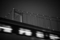B&W photo of a passenger train rushing by in the foreground with a bridge in the background at night.
