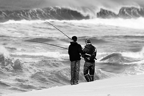 A father and son fish from the shore while huge winter surf breaks off shore. 
