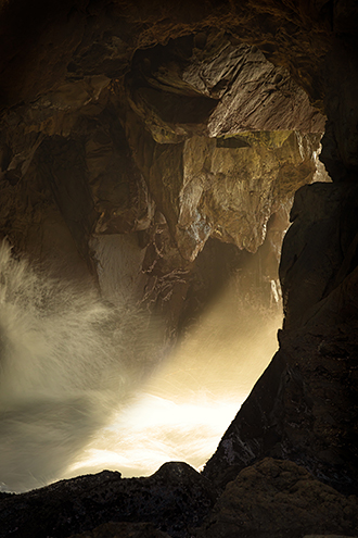 A beam of late afternoon light pushes through the ocean mist of breaking waves in a sea cave. 